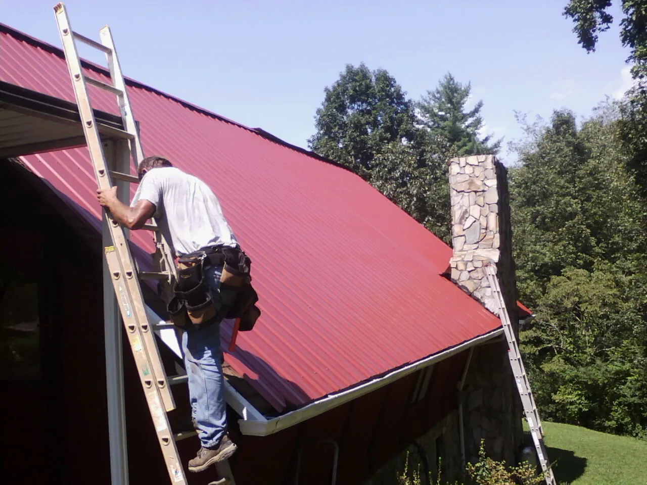 Roofer on a ladder beside a red metal roof and stone chimney — Soffit & Fascia Repair by Roofing Brothers in Fords